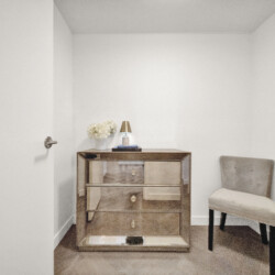 Mirrored three-drawer dresser in a small bedroom corner, topped with white flowers, a brass lamp, and books; a beige upholstered chair nearby.