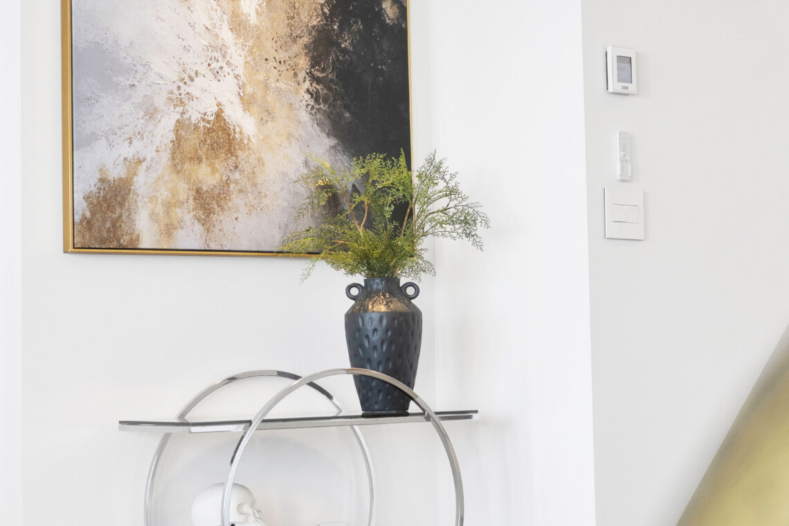 Abstract gold-black artwork in a gold frame on a white wall above a glass-and-metal console with a blue vase of greenery and decorative skulls/books inset below.