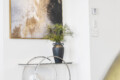 Abstract gold-black artwork in a gold frame on a white wall above a glass-and-metal console with a blue vase of greenery and decorative skulls/books inset below.