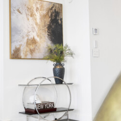 Abstract gold-black artwork in a gold frame on a white wall above a glass-and-metal console with a blue vase of greenery and decorative skulls/books inset below.