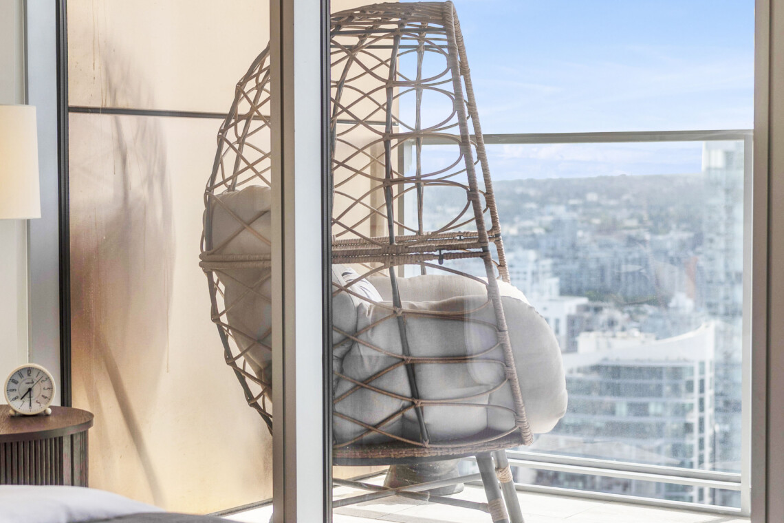 Bedroom scene with a woven wicker chair by glass doors, cushions in the chair, and a city skyline visible outside on a sunny day.