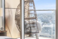 Bedroom scene with a woven wicker chair by glass doors, cushions in the chair, and a city skyline visible outside on a sunny day.