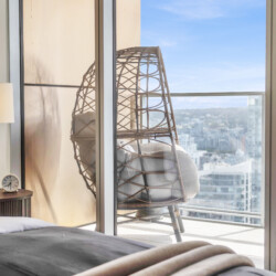 Bedroom scene with a woven wicker chair by glass doors, cushions in the chair, and a city skyline visible outside on a sunny day.