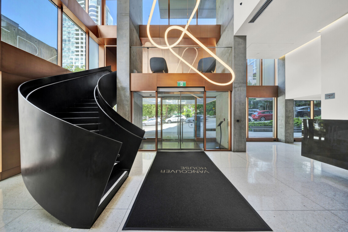 Modern lobby with a large black curved staircase on the left, copper panels, glass doors, and a looping neon sculpture overhead.