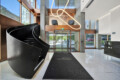 Modern lobby with a large black curved staircase on the left, copper panels, glass doors, and a looping neon sculpture overhead.