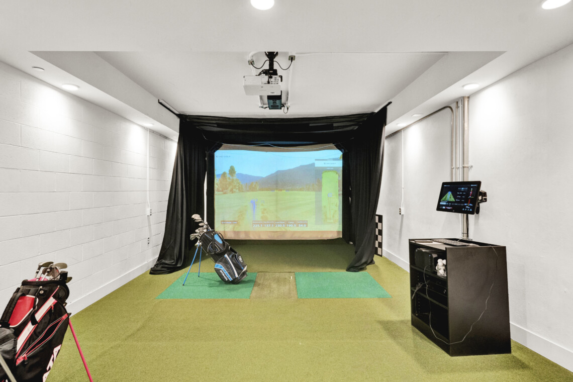 Indoor golf simulator bay with a projection screen, hitting mats, and golf bags along the left wall