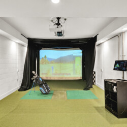Indoor golf simulator bay with a projection screen, hitting mats, and golf bags along the left wall