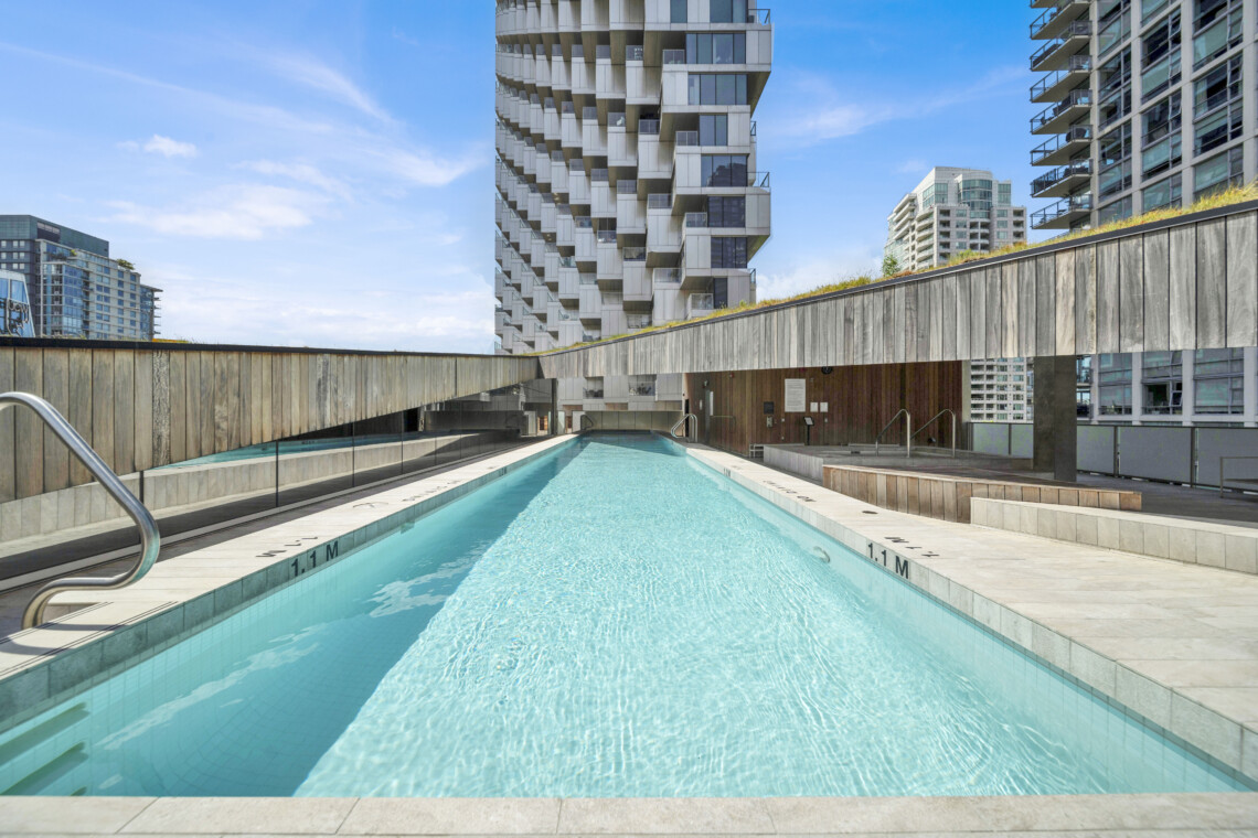 Rooftop rectangular pool surrounded by modern high-rise buildings under a blue sky, with wooden railing and deck area nearby.