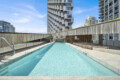 Rooftop rectangular pool surrounded by modern high-rise buildings under a blue sky, with wooden railing and deck area nearby.