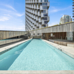 Rooftop rectangular pool surrounded by modern high-rise buildings under a blue sky, with wooden railing and deck area nearby.