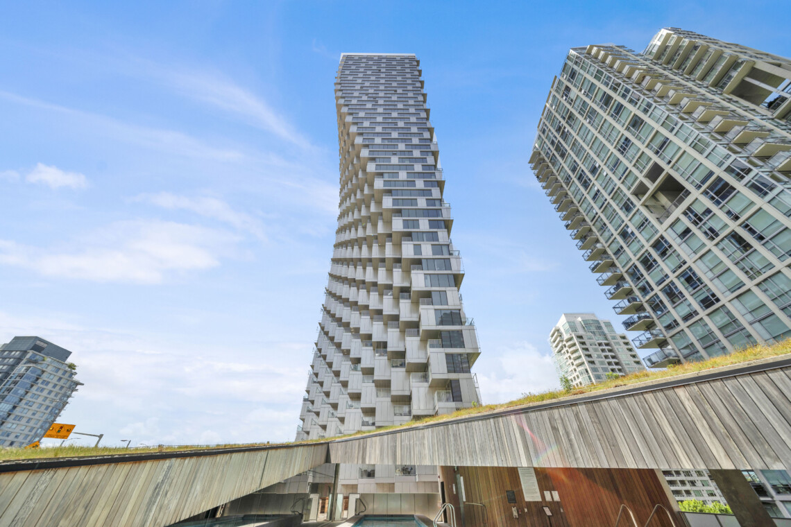 Two modern high-rise buildings with staggered balconies rise against a clear blue sky, featuring glass façades and white panels.