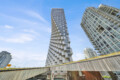 Two modern high-rise buildings with staggered balconies rise against a clear blue sky, featuring glass façades and white panels.