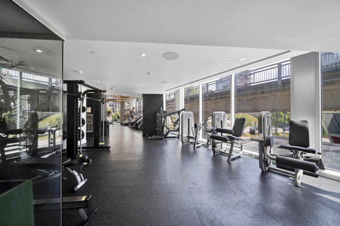 Well-lit gym with cardio and strength machines along large glass walls and a city view outside.