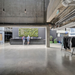 Industrial lobby reception with concrete ceiling, exposed pipes, and a vertical garden behind a dark desk where three people stand talking.