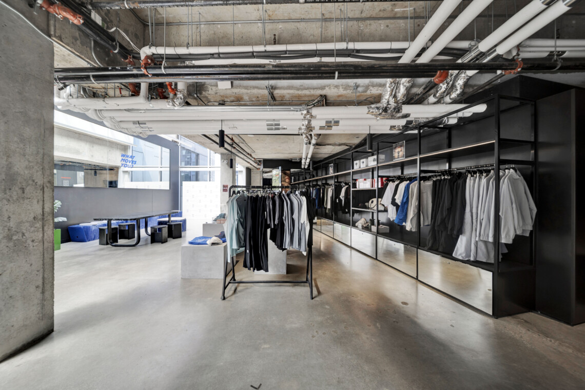 Industrial-style clothing store interior with metal racks of shirts along a glass wall and exposed pipes overhead, giving an urban warehouse vibe.