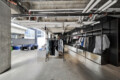 Industrial-style clothing store interior with metal racks of shirts along a glass wall and exposed pipes overhead, giving an urban warehouse vibe.
