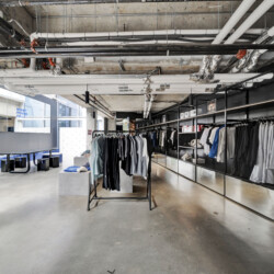 Industrial-style clothing store interior with metal racks of shirts along a glass wall and exposed pipes overhead, giving an urban warehouse vibe.