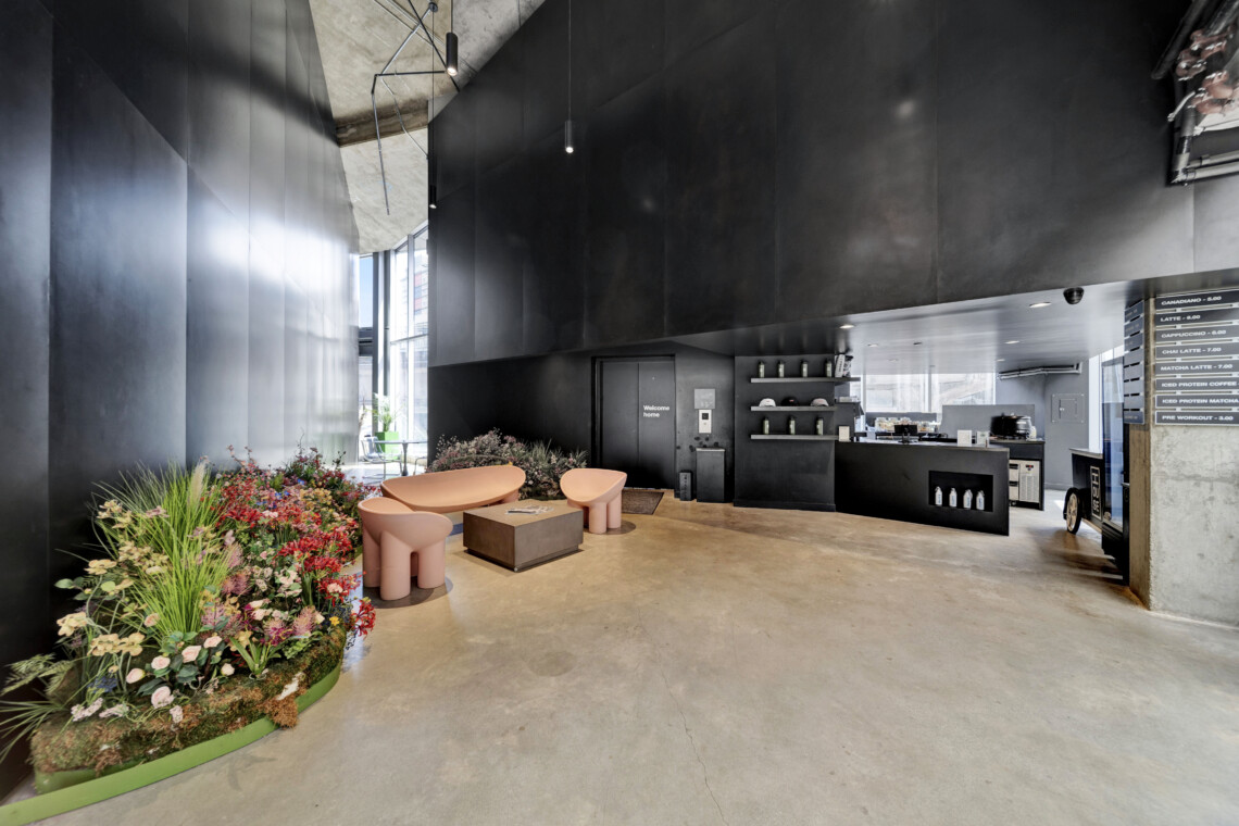 Industrial lobby with black walls and concrete ceiling; pink curved seating around a square table, and a colorful flower planter to the left; coffee bar on the right.