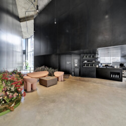 Industrial lobby with black walls and concrete ceiling; pink curved seating around a square table, and a colorful flower planter to the left; coffee bar on the right.
