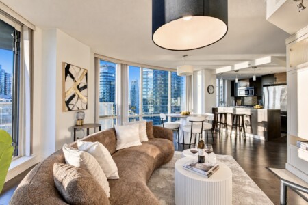 Open-concept living room with curved floor-to-ceiling windows overlooking a city skyline, brown sofa, and white coffee table.