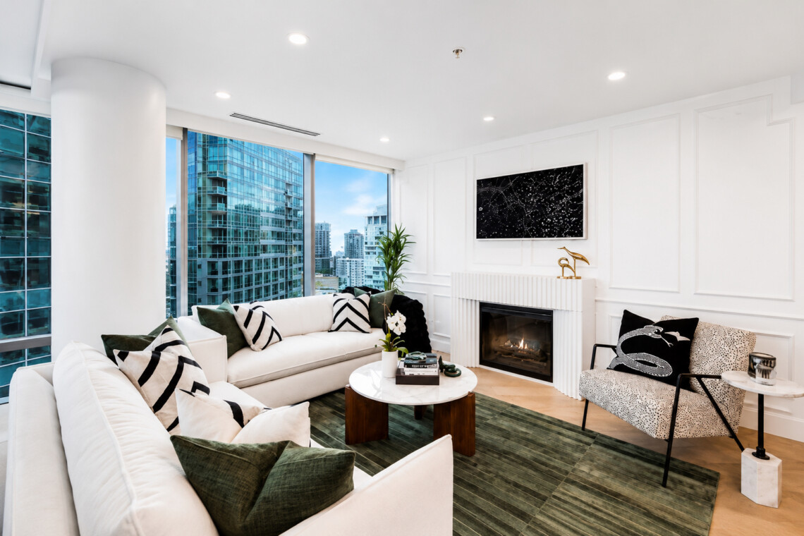 Modern living room with a white sectional, green cushions, a round marble coffee table, and a fireplace, seen from a cozy seating area with a city view.