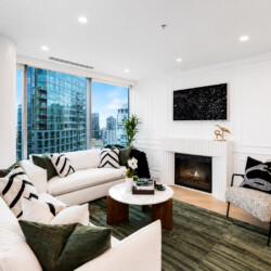 Modern living room with a white sectional, green cushions, a round marble coffee table, and a fireplace, seen from a cozy seating area with a city view.
