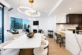 Open-concept living area with marble kitchen counter, circular gold pendant lights, and a beige sofa facing large city windows.