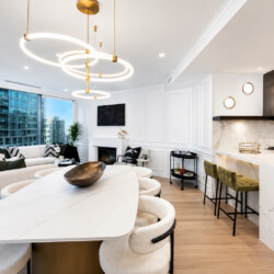 Open-concept living area with marble kitchen counter, circular gold pendant lights, and a beige sofa facing large city windows.