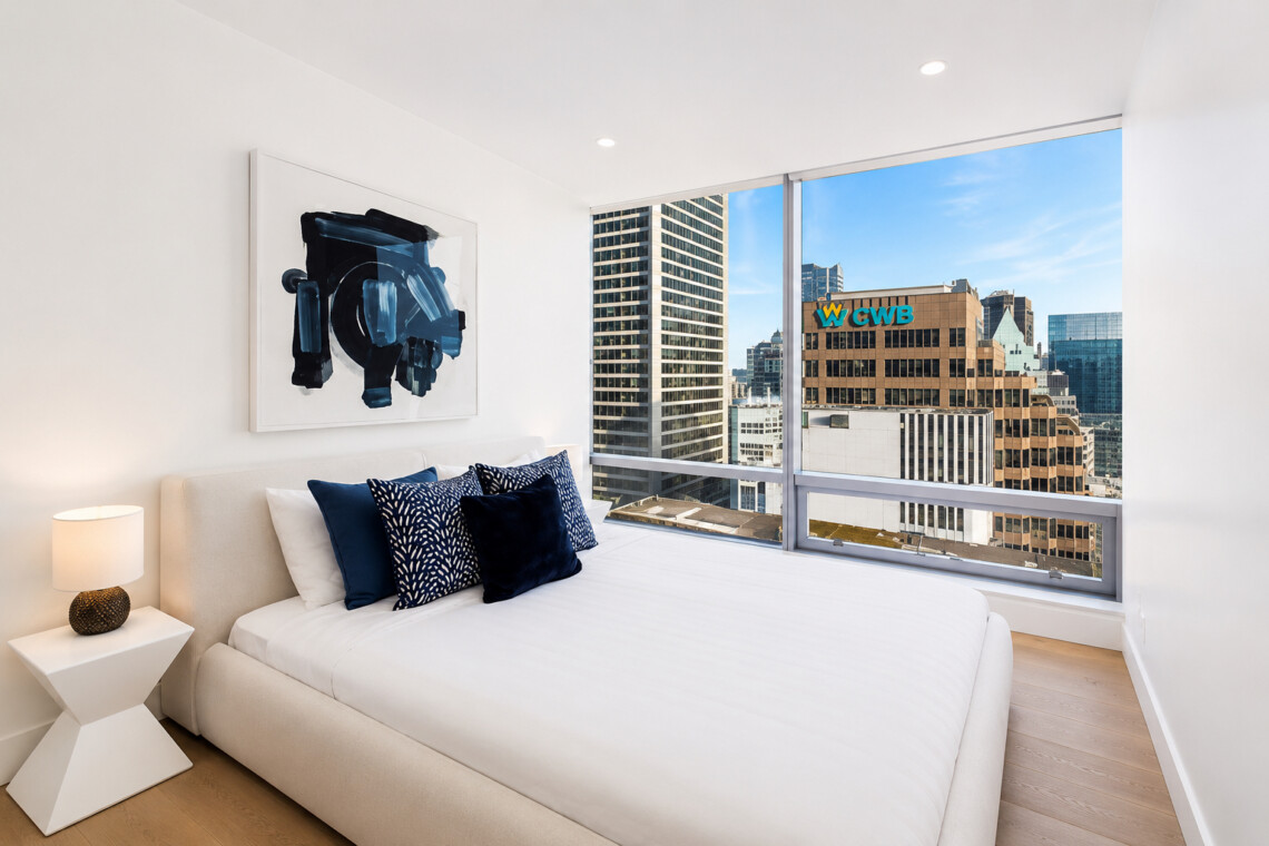 Modern bedroom with white bed, navy pillows, abstract blue art, and a city skyline view through large windows.