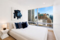 Modern bedroom with white bed, navy pillows, abstract blue art, and a city skyline view through large windows.