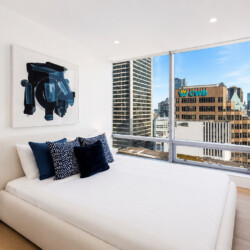 Modern bedroom with white bed, navy pillows, abstract blue art, and a city skyline view through large windows.