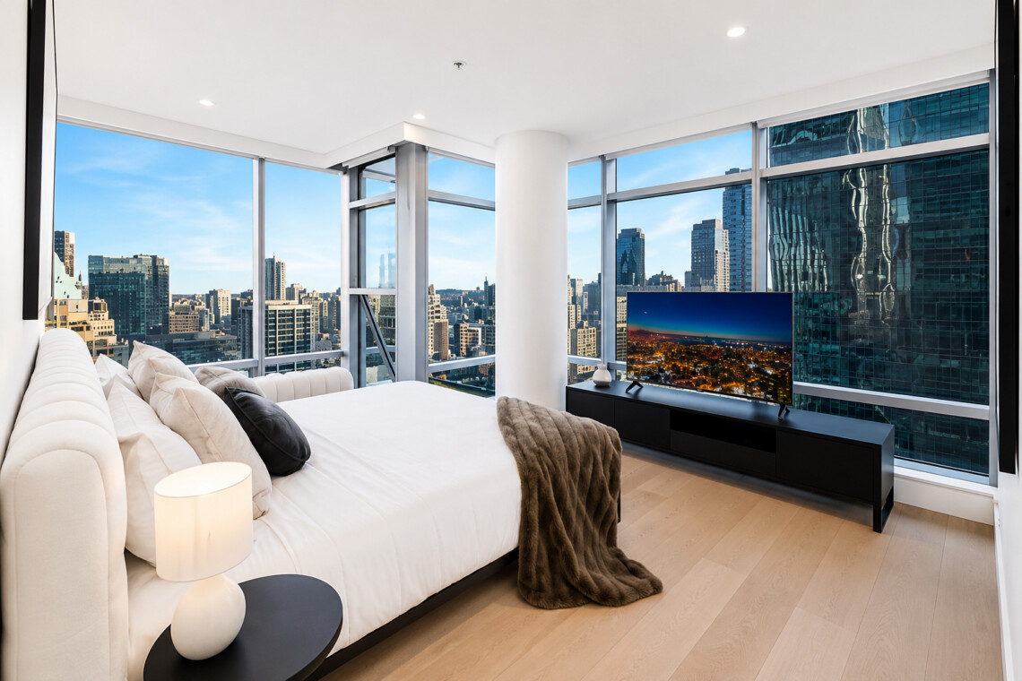 Modern bedroom with white bedding and a fur throw, large windows showing a city skyline outside.