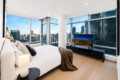 Modern bedroom with white bedding and a fur throw, large windows showing a city skyline outside.