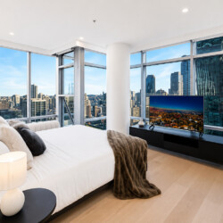 Modern bedroom with white bedding and a fur throw, large windows showing a city skyline outside.