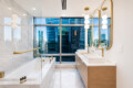 Luxurious bathroom with marble surfaces, gold fixtures, double sinks, and a city skyline visible through large windows behind the vanity.