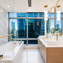 Luxurious bathroom with marble surfaces, gold fixtures, double sinks, and a city skyline visible through large windows behind the vanity.