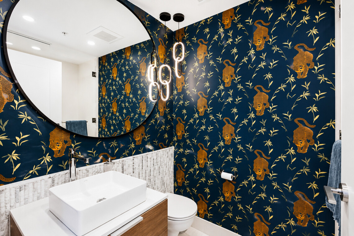 Round mirror above a white rectangular sink in a bathroom with dark blue wallpaper featuring gold leopards and bamboo, and warm pendant lights.