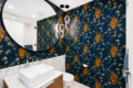 Round mirror above a white rectangular sink in a bathroom with dark blue wallpaper featuring gold leopards and bamboo, and warm pendant lights.