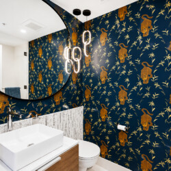 Round mirror above a white rectangular sink in a bathroom with dark blue wallpaper featuring gold leopards and bamboo, and warm pendant lights.