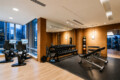 A modern gym with cardio machines by large windows overlooking a city skyline, wood-paneled walls and mirrored wall.