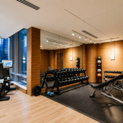 A modern gym with cardio machines by large windows overlooking a city skyline, wood-paneled walls and mirrored wall.