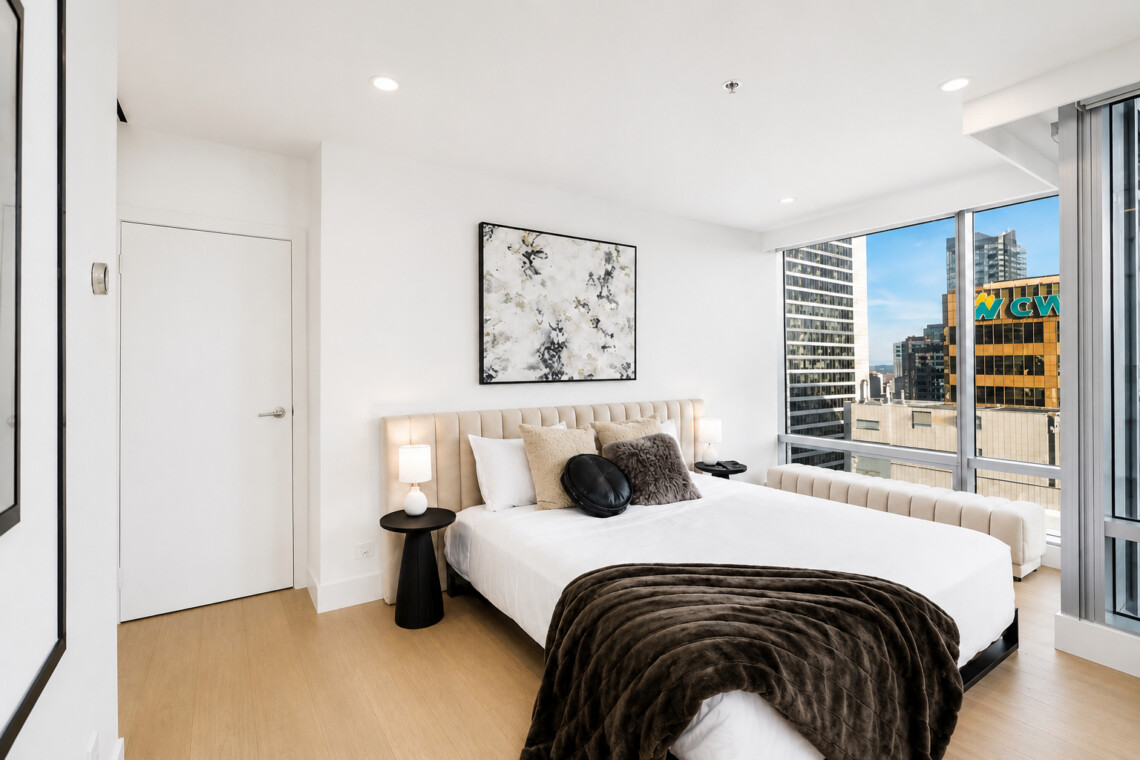 Modern bedroom with a beige tufted headboard bed, white linens, brown fur throw, and abstract wall art, plus a city view through large windows.