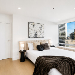Modern bedroom with a beige tufted headboard bed, white linens, brown fur throw, and abstract wall art, plus a city view through large windows.