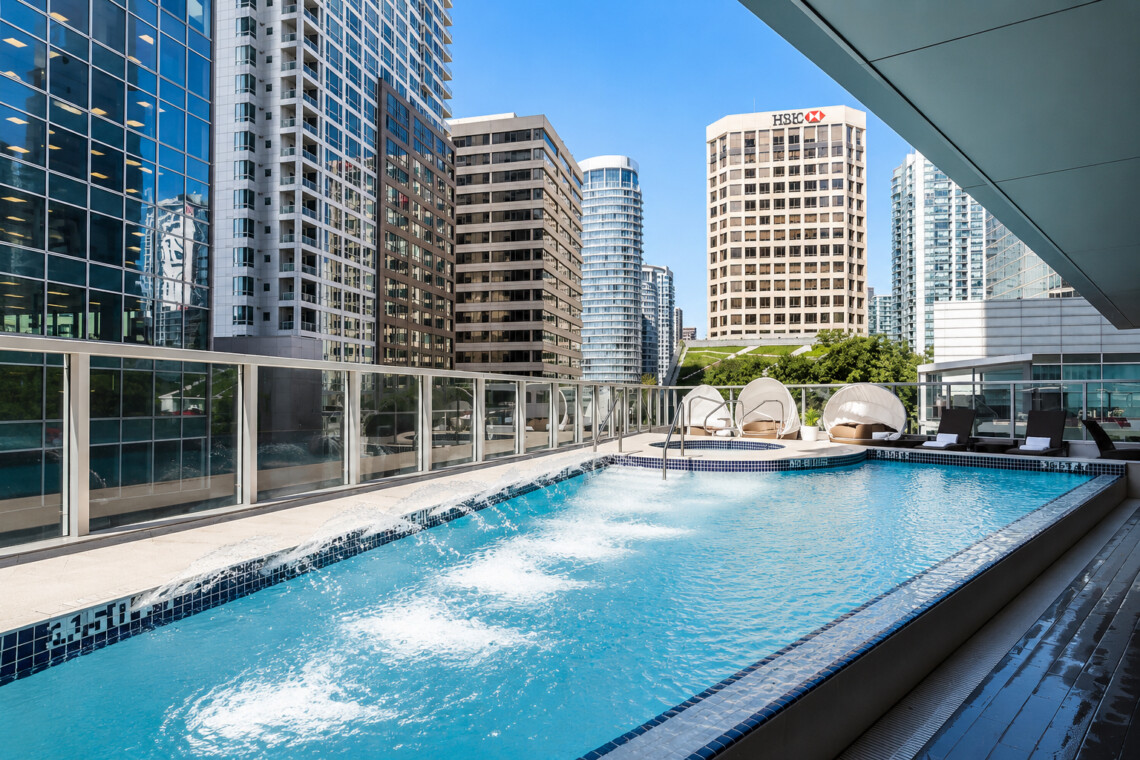 Rooftop pool with loungers and cabanas, surrounded by glass-walled skyscrapers against a blue sky