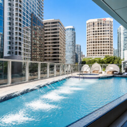 Rooftop pool with loungers and cabanas, surrounded by glass-walled skyscrapers against a blue sky