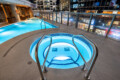 Rooftop pool and circular illuminated hot tub on a hotel terrace at night, with glass railing and a city skyline in the background.