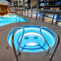 Rooftop pool and circular illuminated hot tub on a hotel terrace at night, with glass railing and a city skyline in the background.