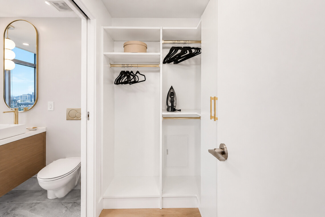 Modern bathroom with a storage closet containing hanging rods, hangers, shelves, and a small iron on the middle shelf, adjacent to a white vanity and toilet.