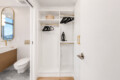 Modern bathroom with a storage closet containing hanging rods, hangers, shelves, and a small iron on the middle shelf, adjacent to a white vanity and toilet.