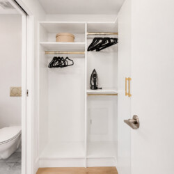Modern bathroom with a storage closet containing hanging rods, hangers, shelves, and a small iron on the middle shelf, adjacent to a white vanity and toilet.